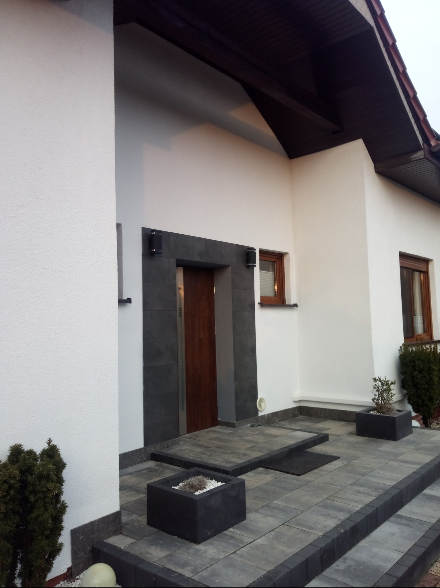 Nowoczesny dom jednorodzinny z jasną elewacją, ciemną stolarką okienną i wejściem wyłożonym szarą kostką brukową, z donicami z roślinami.