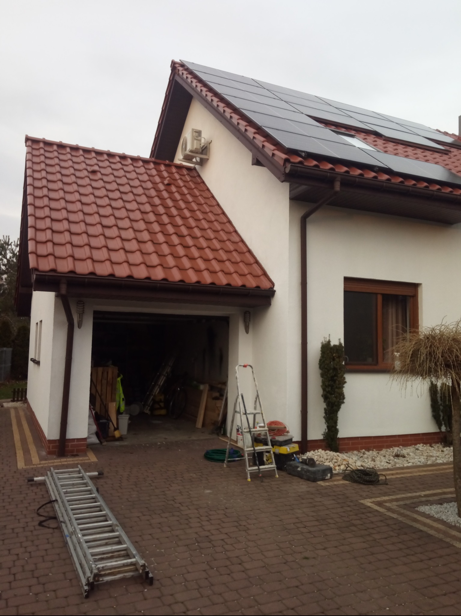 Dom jednorodzinny z białymi ścianami, czerwoną dachówką i panelami słonecznymi na dachu, drabina oparta o ścianę, otwarty garaż z narzędziami w środku, kostka brukowa przed domem.
