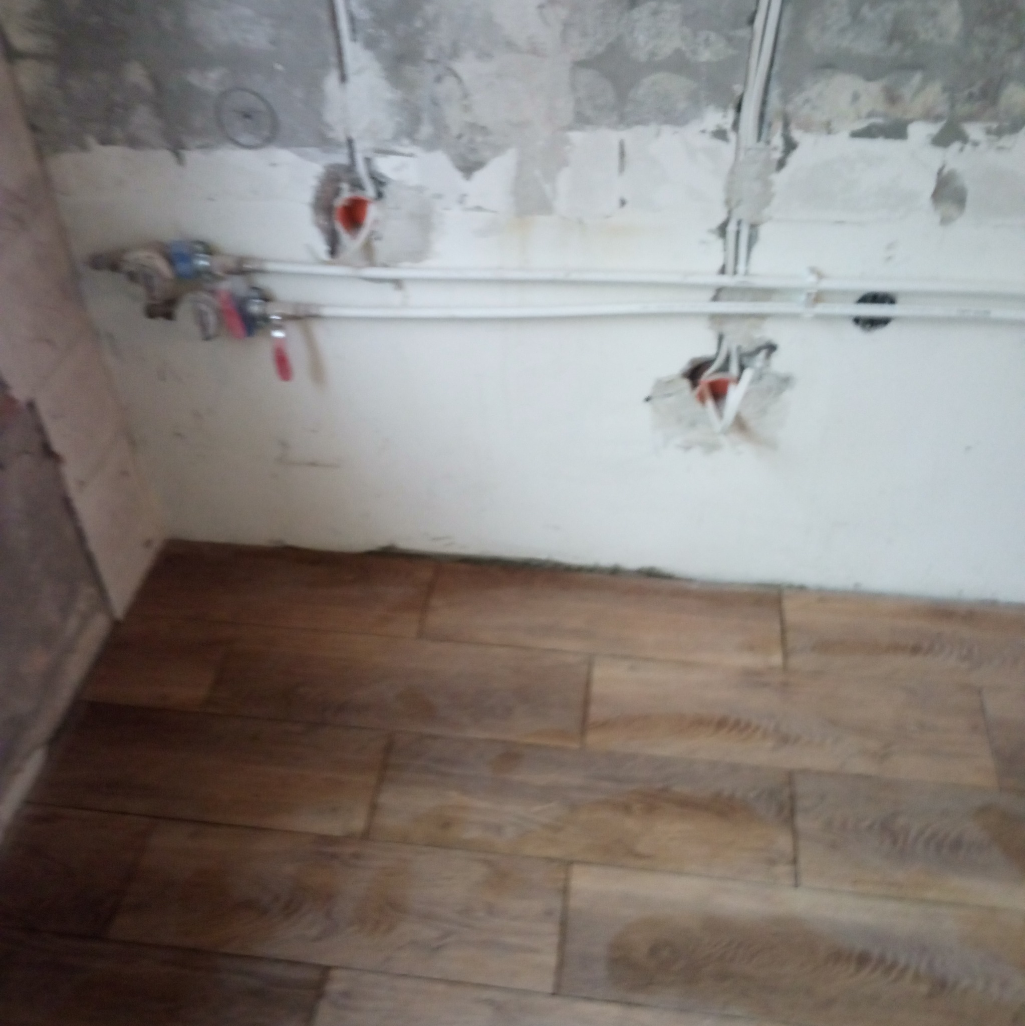 Ułożona podłoga z paneli imitujących drewno, widoczne instalacje wodne i elektryczne w surowej ścianie w trakcie remontu.