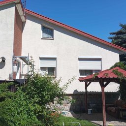 usługi ogólnobudowlane u Ilony - Dom jednorodzinny z białą elewacją, czerwonym dachem i roletami zewnętrznymi; widoczne rusztowanie, fragment ceglanej ściany i altana z zarośniętym dachem.