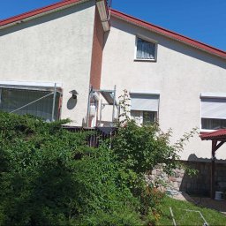 usługi ogólnobudowlane u Ilony - Dwupiętrowy dom z jasną elewacją i czerwoną dachówką, częściowo zasłonięty bujną zielenią, widoczne rusztowania przy oknie na piętrze.