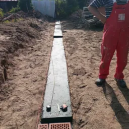 Wykopany i zalany betonem fundament pod ogrodzenie, wytyczony sznurkiem, z widocznymi cegłami klinkierowymi i narzędziami budowlanymi w piaszczystym gruncie.