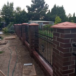 usługi ogólnobudowlane u Ilony - Nowo wybudowane ogrodzenie z czerwonej cegły z czarnymi, kutymi elementami, zakończone czerwonymi daszkami, otoczone zielenią ogrodu.