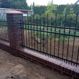 usługi ogólnobudowlane u Ilony - Nowoczesne ogrodzenie z czarnych prętów z dekoracyjnym motywem serca, osadzone na niskim, murowanym cokole z cegły klinkierowej, z filarami z cegły, na tle zieleni ogrodowej.