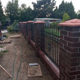 usługi ogólnobudowlane u Ilony - Nowo zbudowane ogrodzenie z czerwonej cegły z czarnymi metalowymi przęsłami dekoracyjnymi, widoczne ślady prac budowlanych w postaci piasku, węża ogrodowego i palet w tle