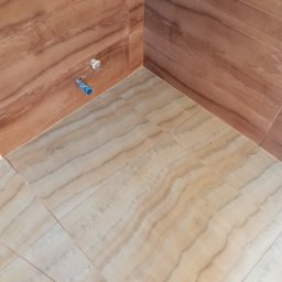 usługi ogólnobudowlane u Ilony - Wykończony narożnik łazienki z brązowymi płytkami ściennymi imitującymi drewno i beżowymi płytkami podłogowymi o wzorze marmuru, widoczne przyłącza hydrauliczne.