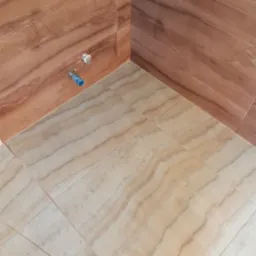 Wykończony narożnik łazienki z brązowymi płytkami ściennymi imitującymi drewno i beżowymi płytkami podłogowymi o wzorze marmuru, widoczne przyłącza hydrauliczne.