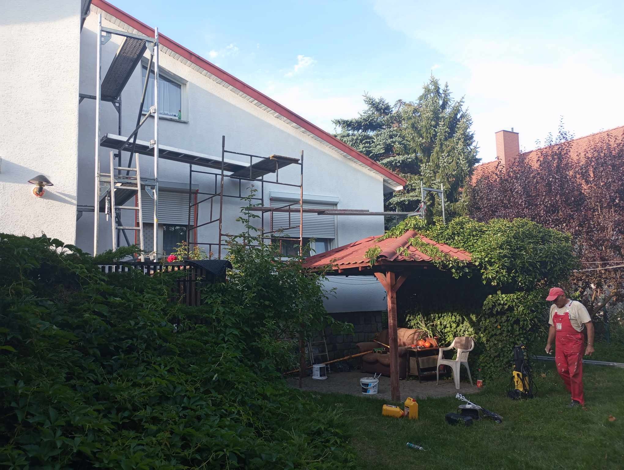Dom jednorodzinny w trakcie malowania elewacji, widoczne rusztowanie, wiaderka z farbą i pracownik w czerwonym kombinezonie na tle bujnej zieleni.