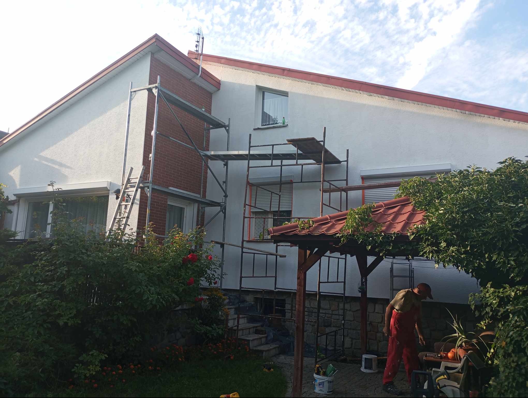 Prace na elewacji domu jednorodzinnego z użyciem rusztowania, widoczny fragment ściany z cegły, częściowo otynkowanej i pomalowanej na biało, pracownik w czerwonym kombinezonie przygotowuje się...