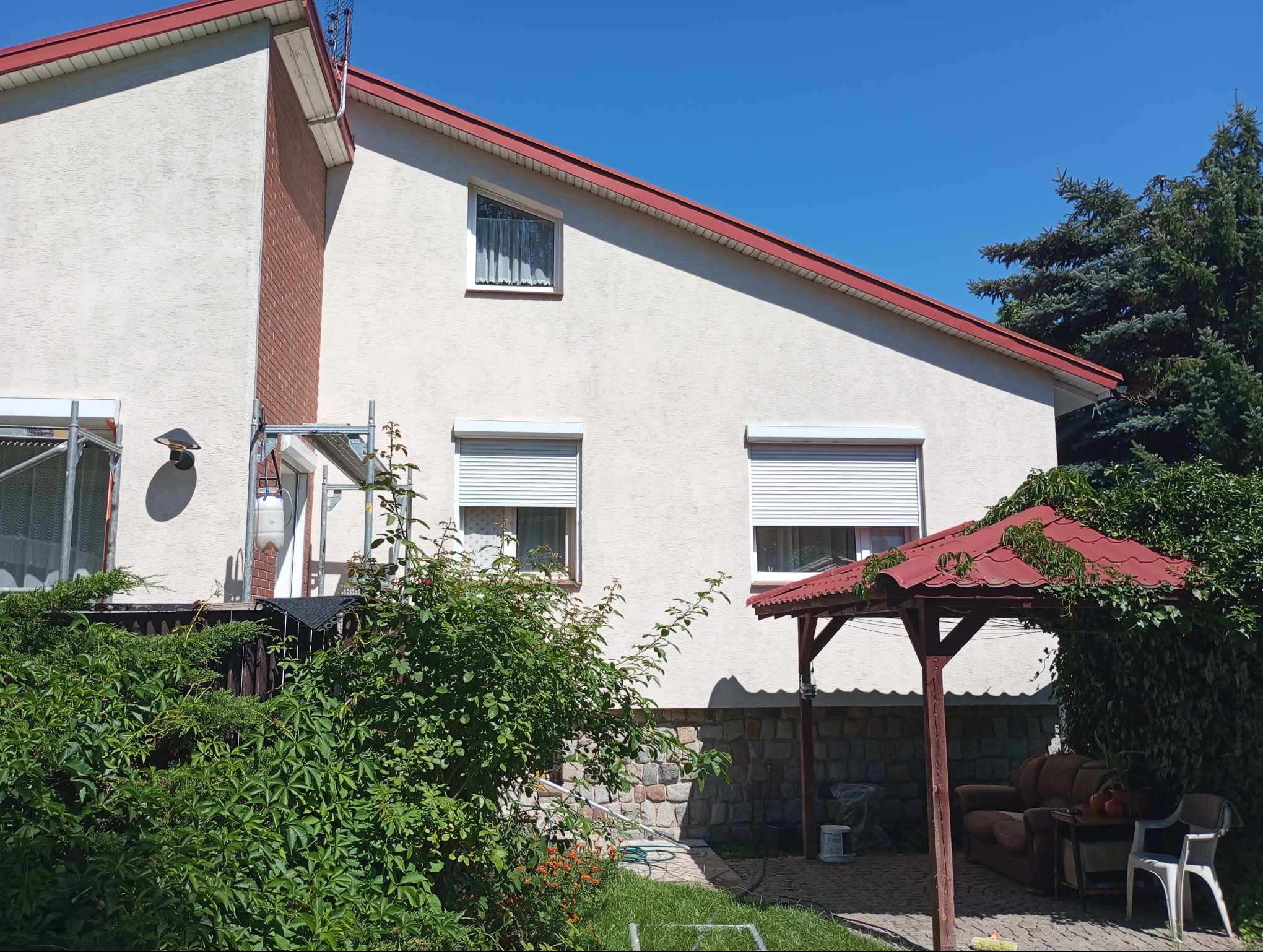 Dom jednorodzinny z białą elewacją, czerwonym dachem i roletami zewnętrznymi; widoczne rusztowanie, fragment ceglanej ściany i altana z zarośniętym dachem.