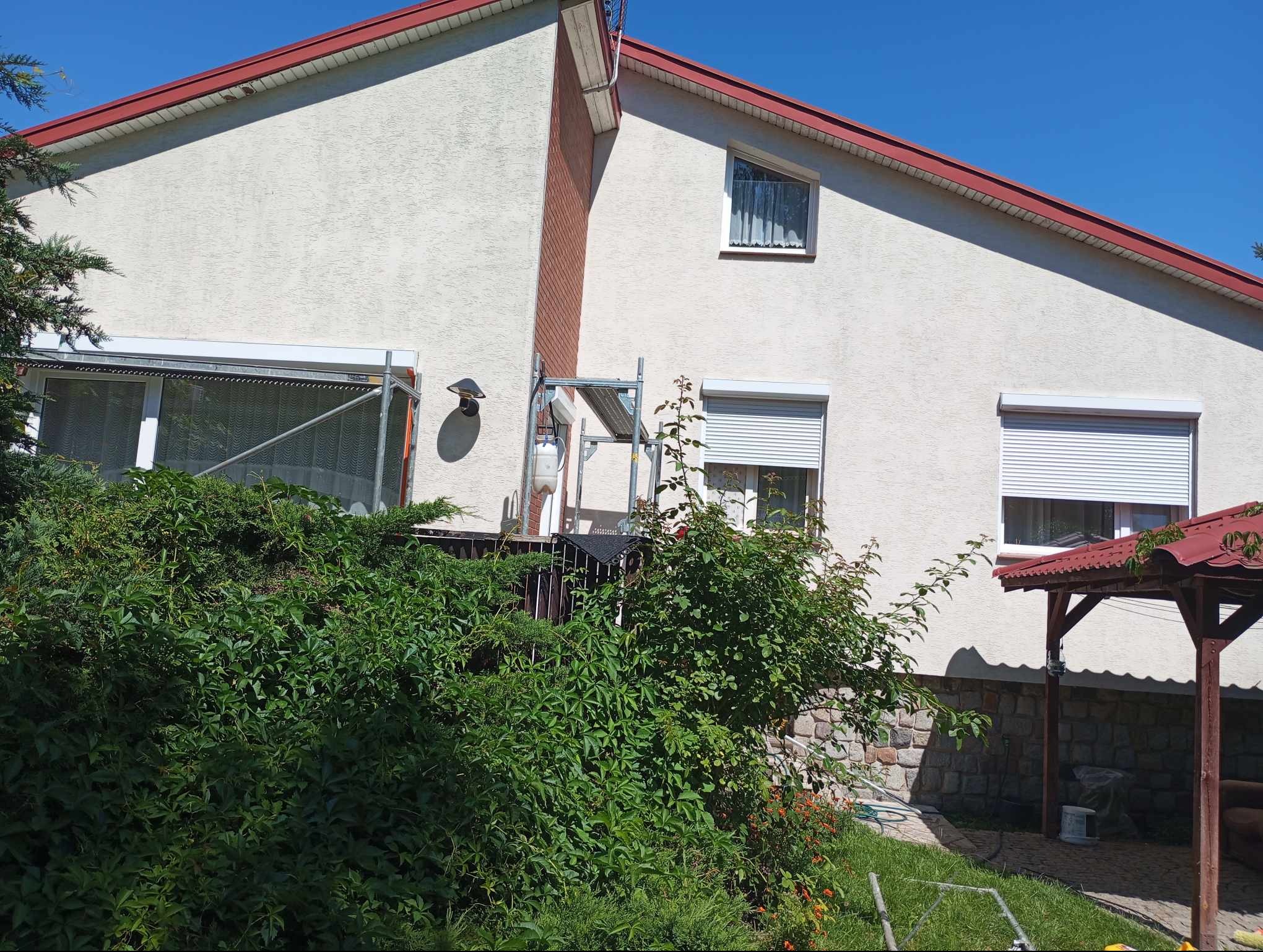 Dwupiętrowy dom z jasną elewacją i czerwoną dachówką, częściowo zasłonięty bujną zielenią, widoczne rusztowania przy oknie na piętrze.