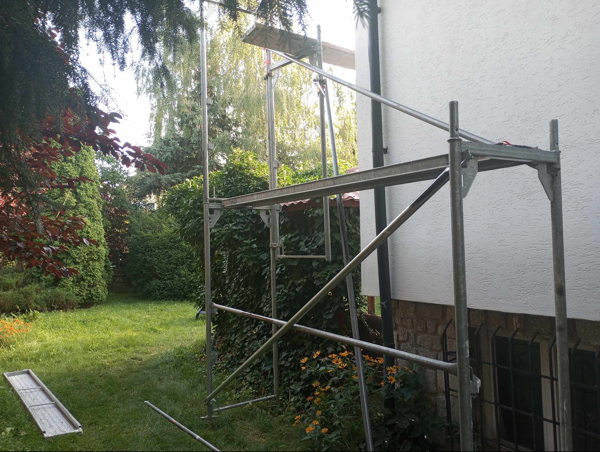Metalowe rusztowanie ustawione przy elewacji domu w Chełmnie, widok z poziomu trawnika z widocznymi kwiatami i elementami zieleni.