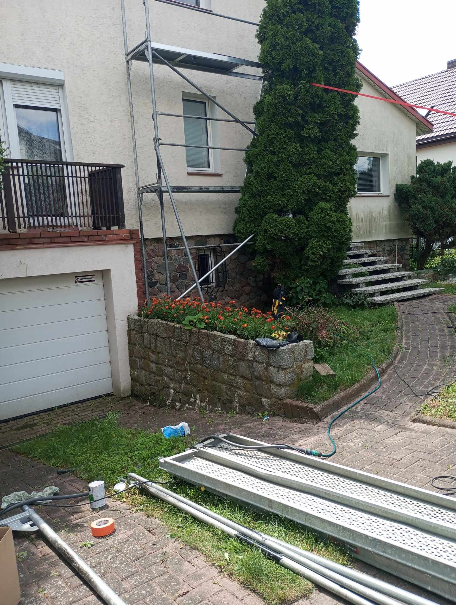 Dom jednorodzinny z elewacją w trakcie przygotowania do malowania, widoczne rusztowanie, teren wokół domu z ogrodem, elementy rusztowania leżące na ziemi.