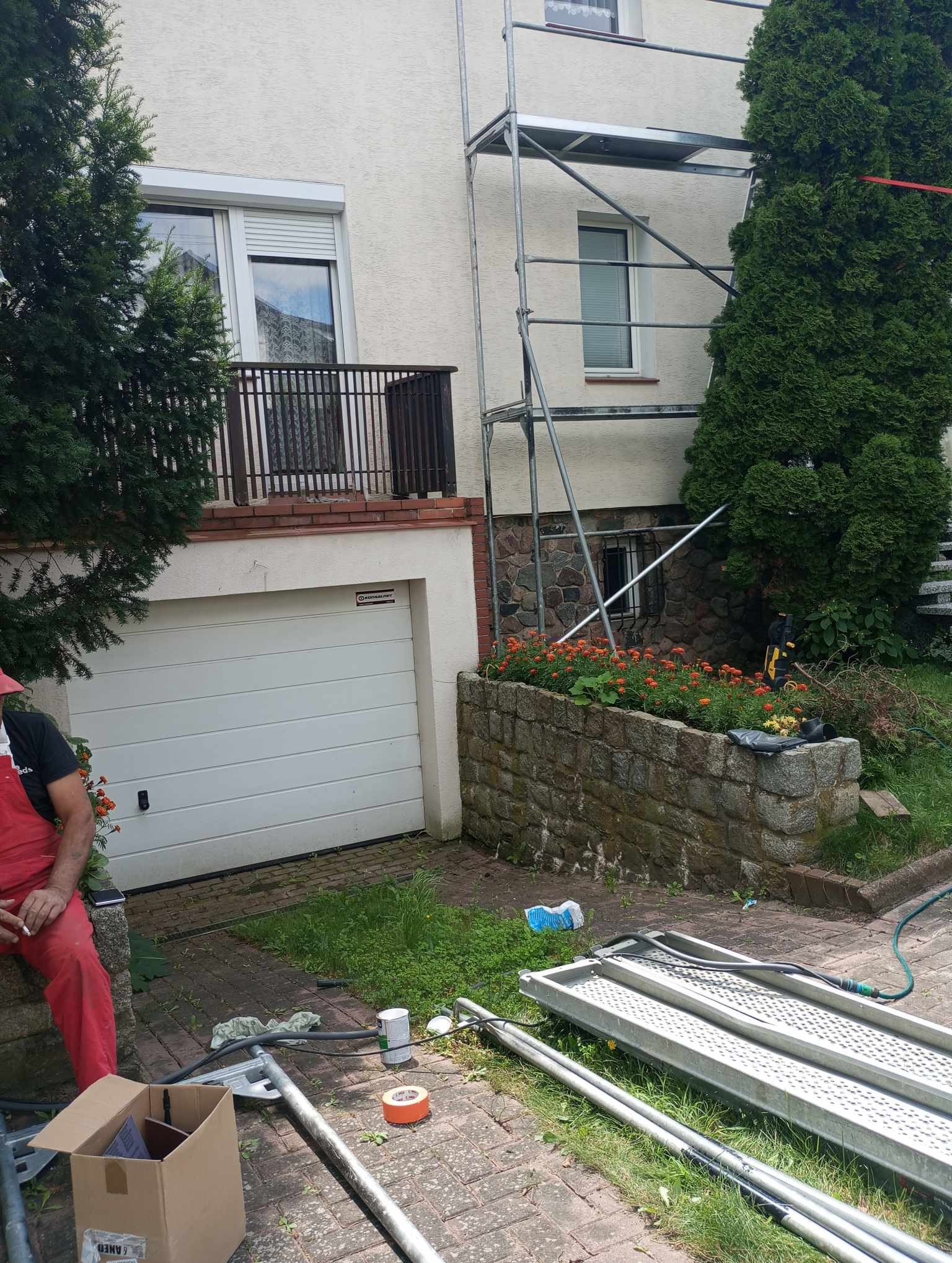 Przygotowania do malowania elewacji: rozstawione rusztowanie, materiały malarskie i pracownik w czerwonym kombinezonie robiący przerwę na papierosa przed budynkiem z garażem i balkonem.