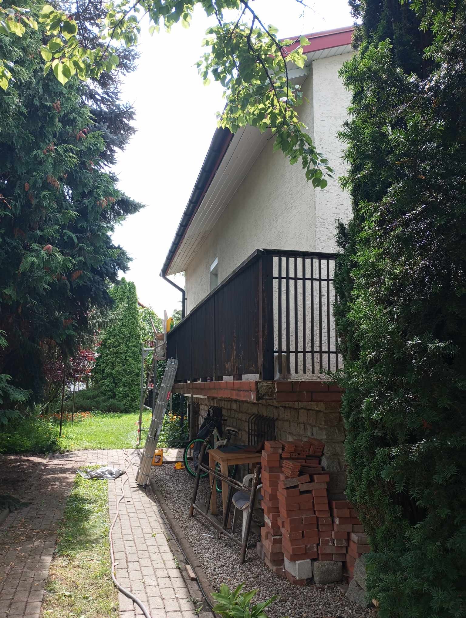 Elewacja domu jednorodzinnego z drewnianą balustradą balkonu, widoczna drabina oparta o ścianę, stos cegieł i narzędzia budowlane w ogrodzie.