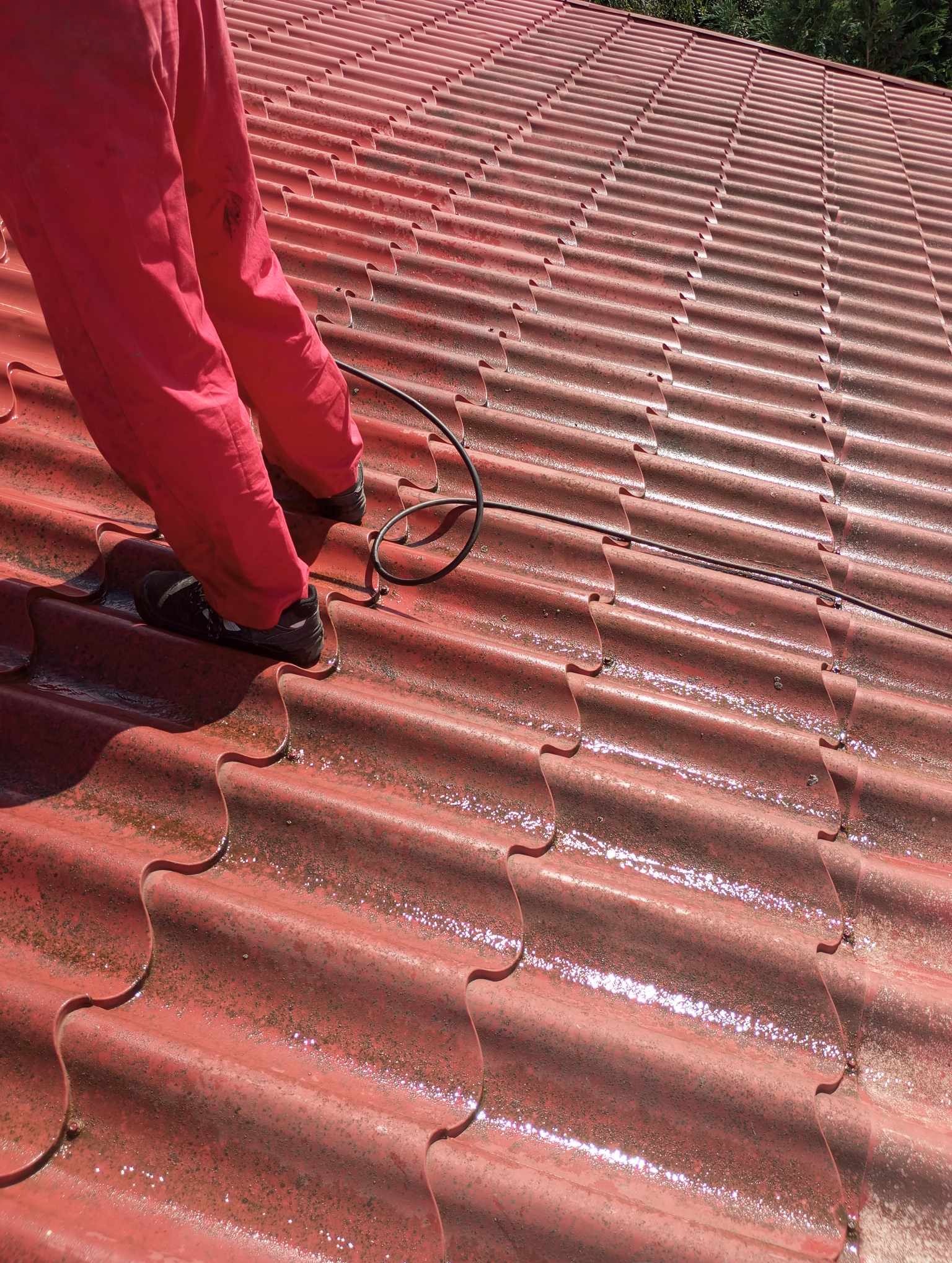 Pracownik w czerwonym kombinezonie stoi na czerwonym, metalowym dachu podczas jego czyszczenia, widoczny kabel ciśnieniowy.