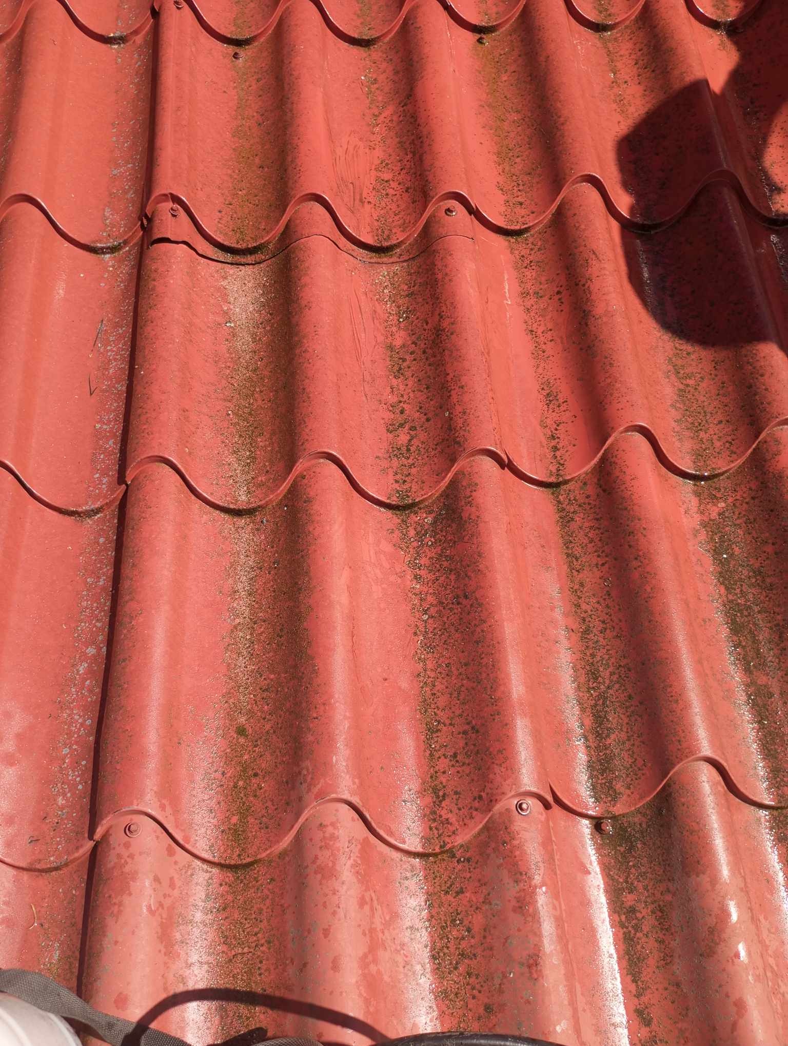 Czerwona, pofałdowana blacha dachowa, pokryta miejscami zielonym nalotem i zabrudzeniami, widoczna z góry pod ostrym kątem.