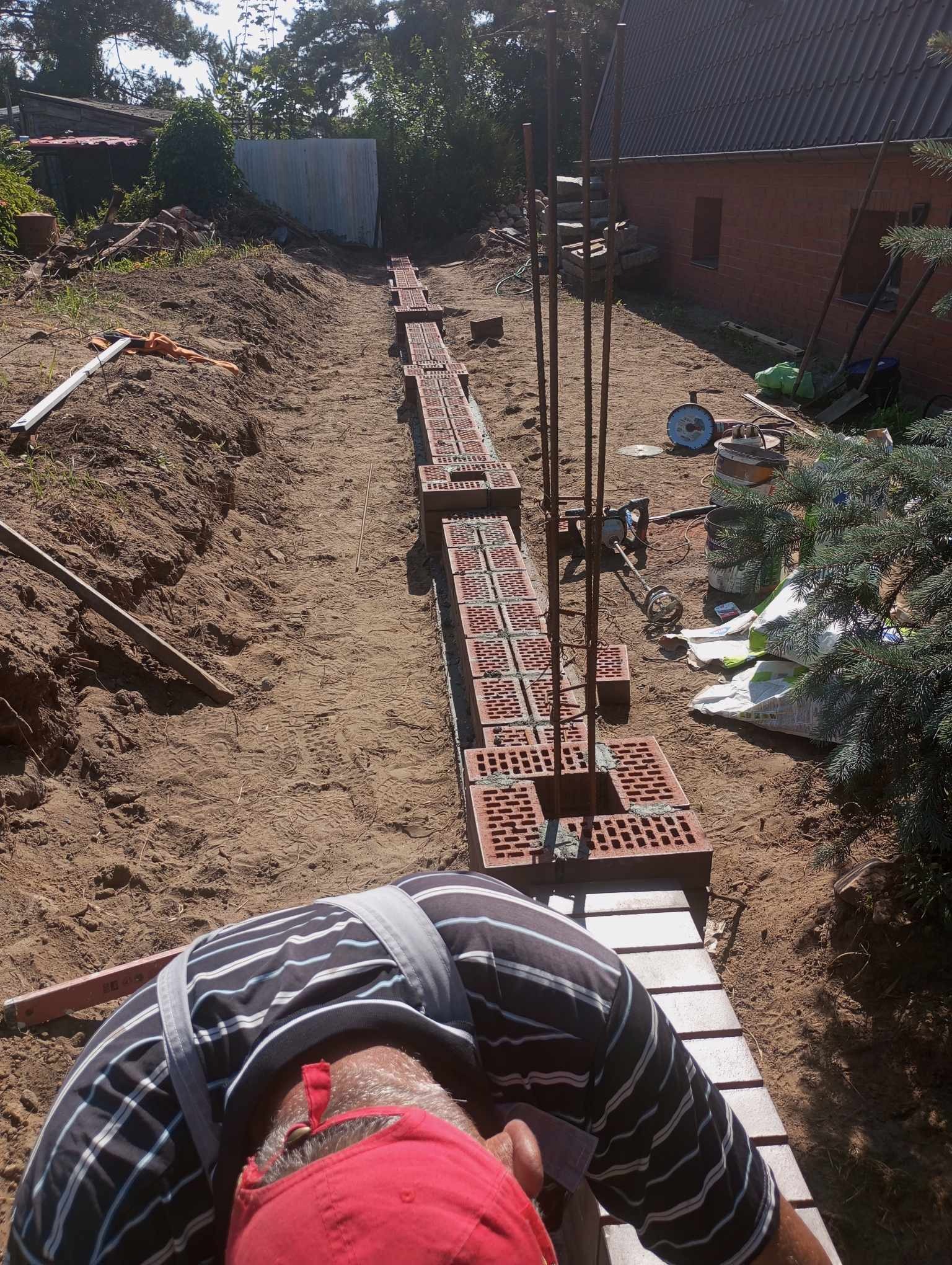 Budowa fundamentu z czerwonej cegły dziurawki z wystającymi prętami zbrojeniowymi, widok z góry na pracownika w pasiastej koszuli i czerwonej czapce, w tle ziemia, narzędzia i budynek z czerwonej...