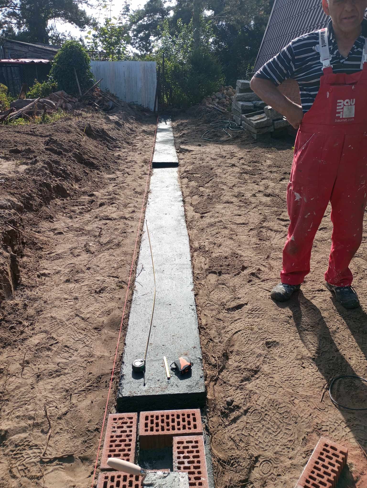 Wykopany i zalany betonem fundament pod ogrodzenie, wytyczony sznurkiem, z widocznymi cegłami klinkierowymi i narzędziami budowlanymi w piaszczystym gruncie.