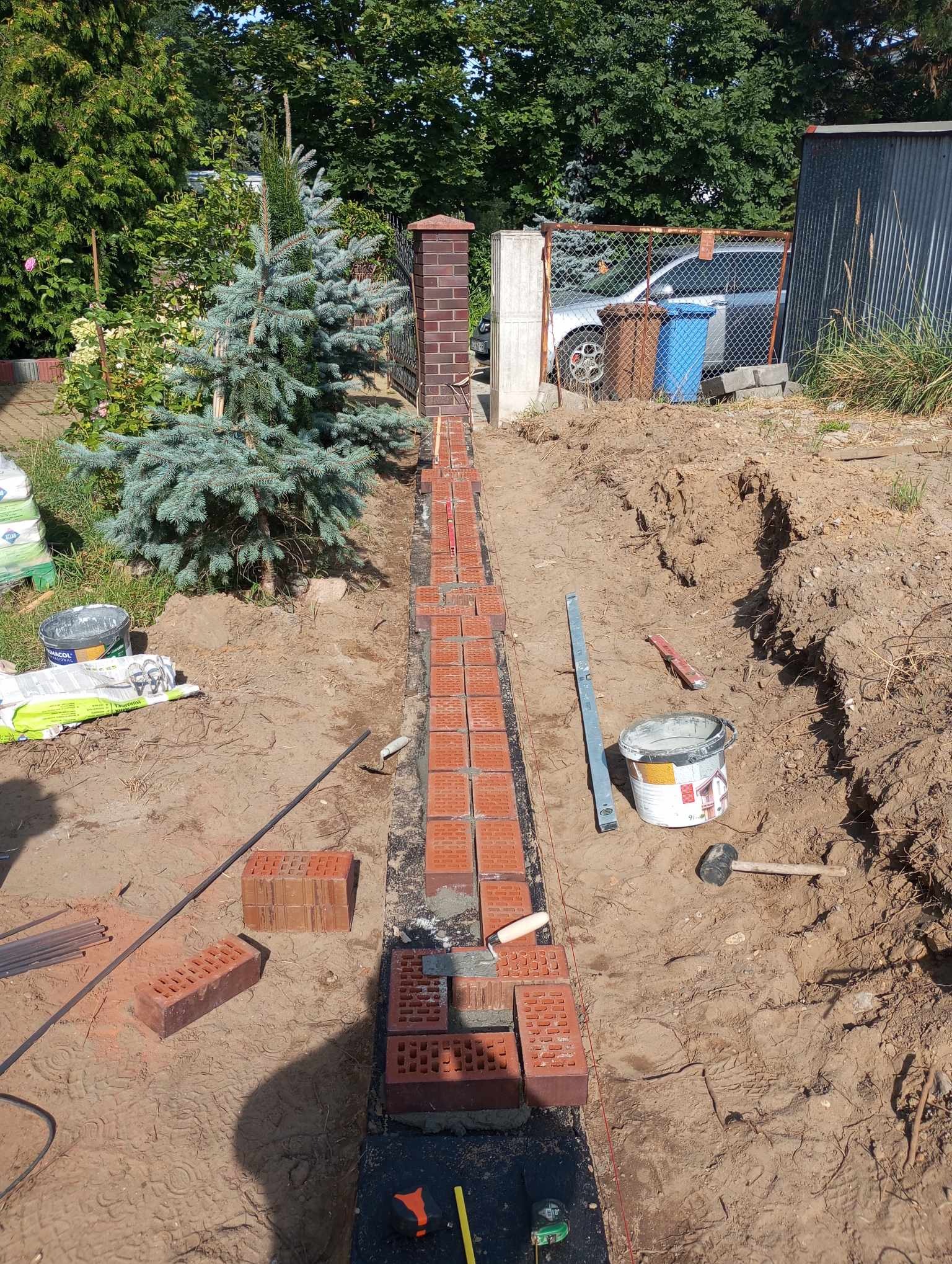 Budowa fundamentu z czerwonej cegły na zewnątrz, widoczne narzędzia, poziomica, sznurek murarski i wiadro z zaprawą.