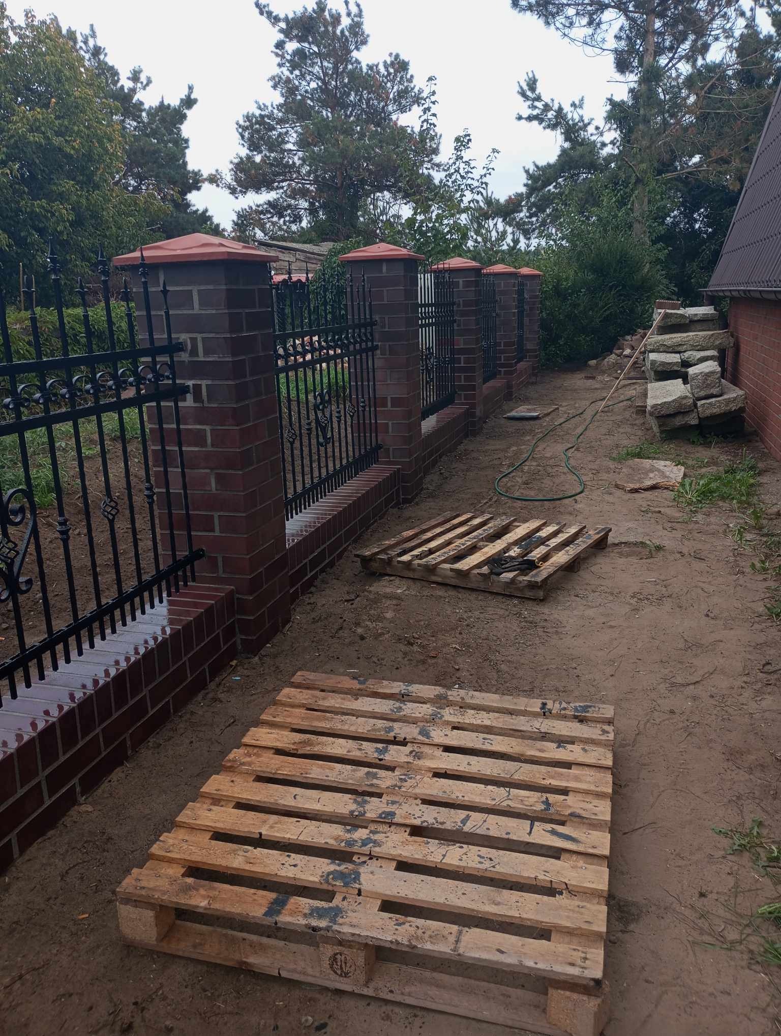 Nowoczesne ogrodzenie z cegły klinkierowej z elementami kutymi w Chełmnie, widoczne palety i stos kamieni sugerują trwające prace.