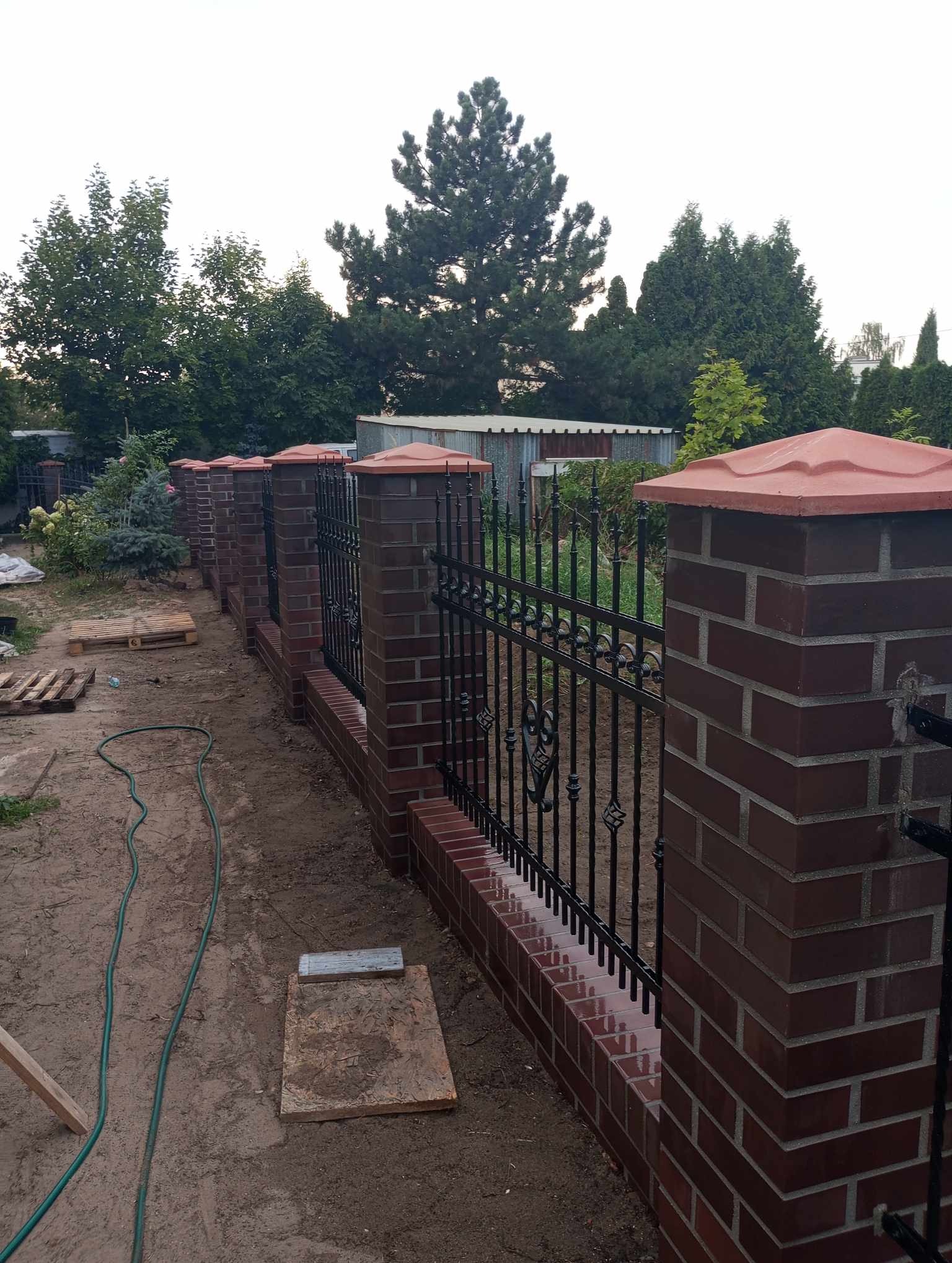 Nowo wybudowane ogrodzenie z czerwonej cegły z czarnymi, kutymi elementami, zakończone czerwonymi daszkami, otoczone zielenią ogrodu.