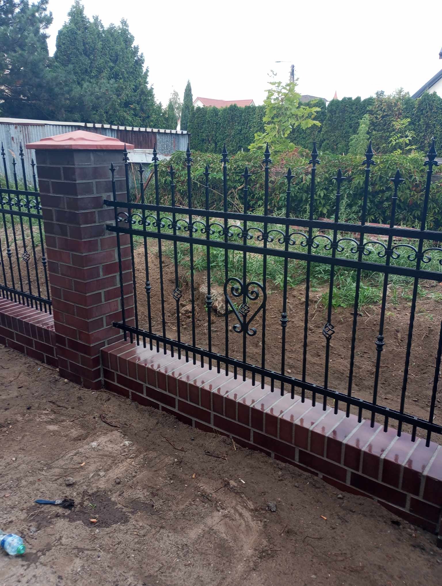 Nowoczesne ogrodzenie z czarnych prętów z dekoracyjnym motywem serca, osadzone na niskim, murowanym cokole z cegły klinkierowej, z filarami z cegły, na tle zieleni ogrodowej.