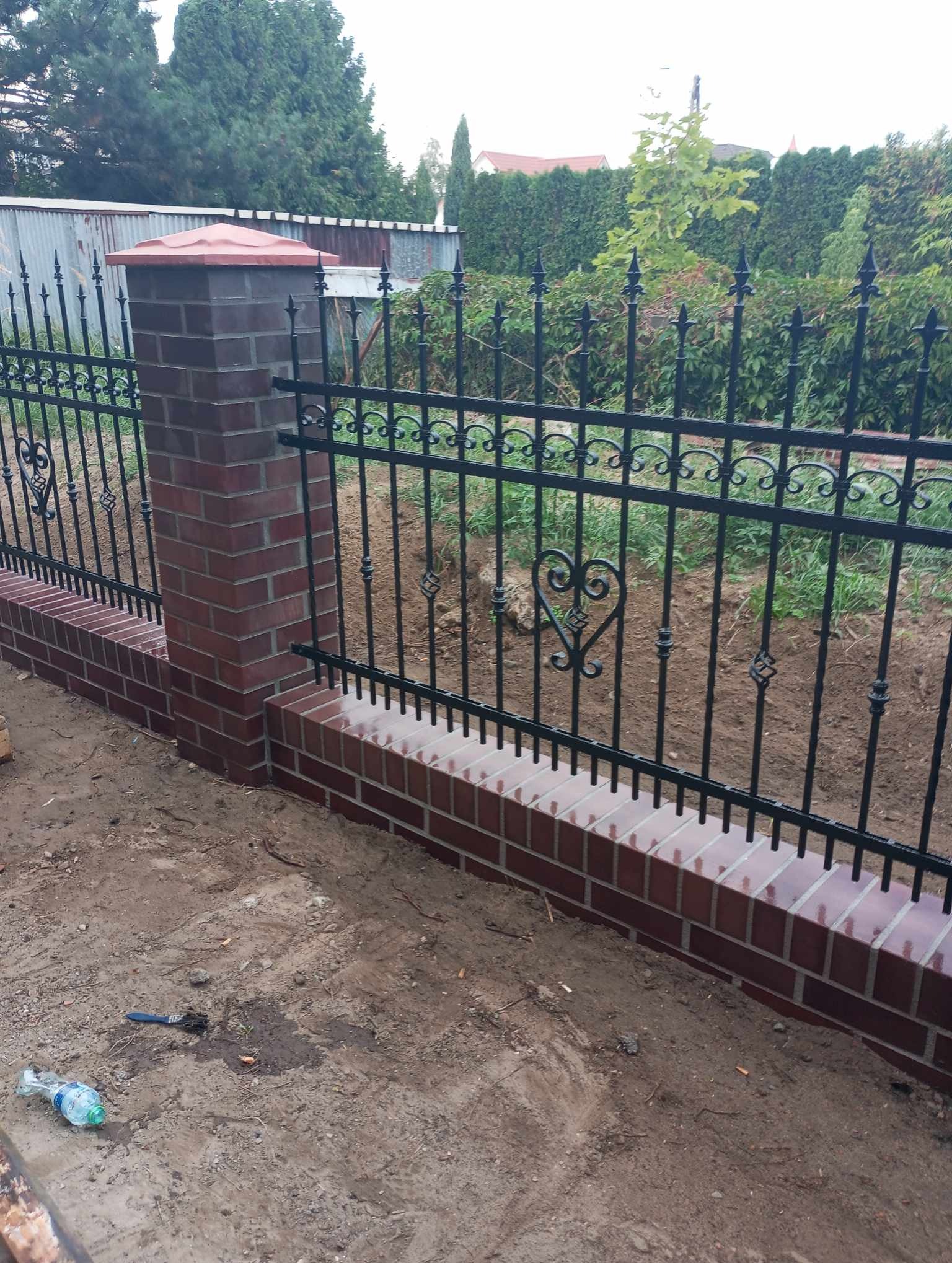 Nowo wybudowane, niskie ogrodzenie z cegły klinkierowej z czarnymi, kutymi przęsłami o dekoracyjnym wzorze serc, widoczne na tle zielonego ogrodu.