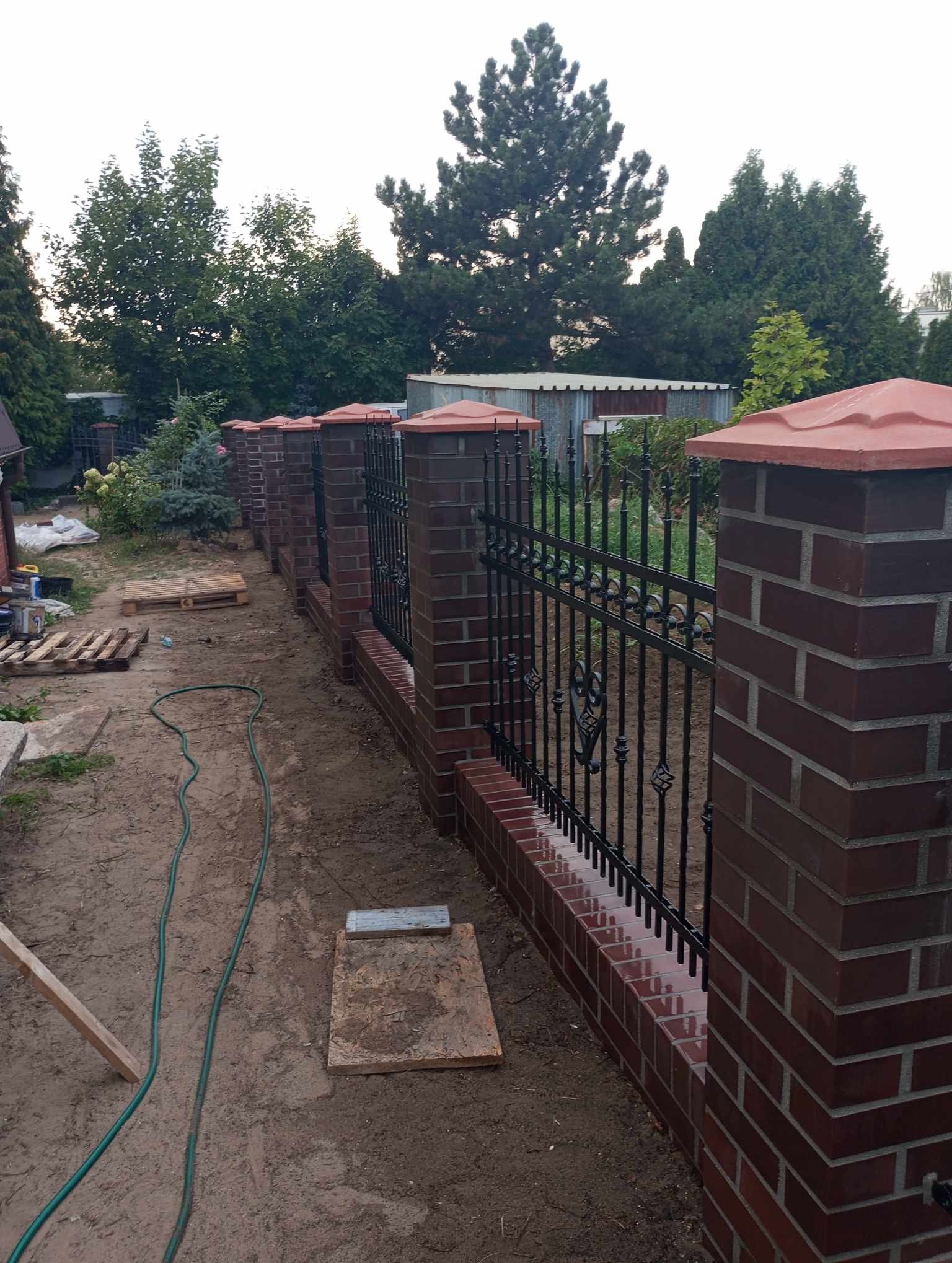 Nowo zbudowane ogrodzenie z czerwonej cegły z czarnymi metalowymi przęsłami dekoracyjnymi, widoczne ślady prac budowlanych w postaci piasku, węża ogrodowego i palet w tle