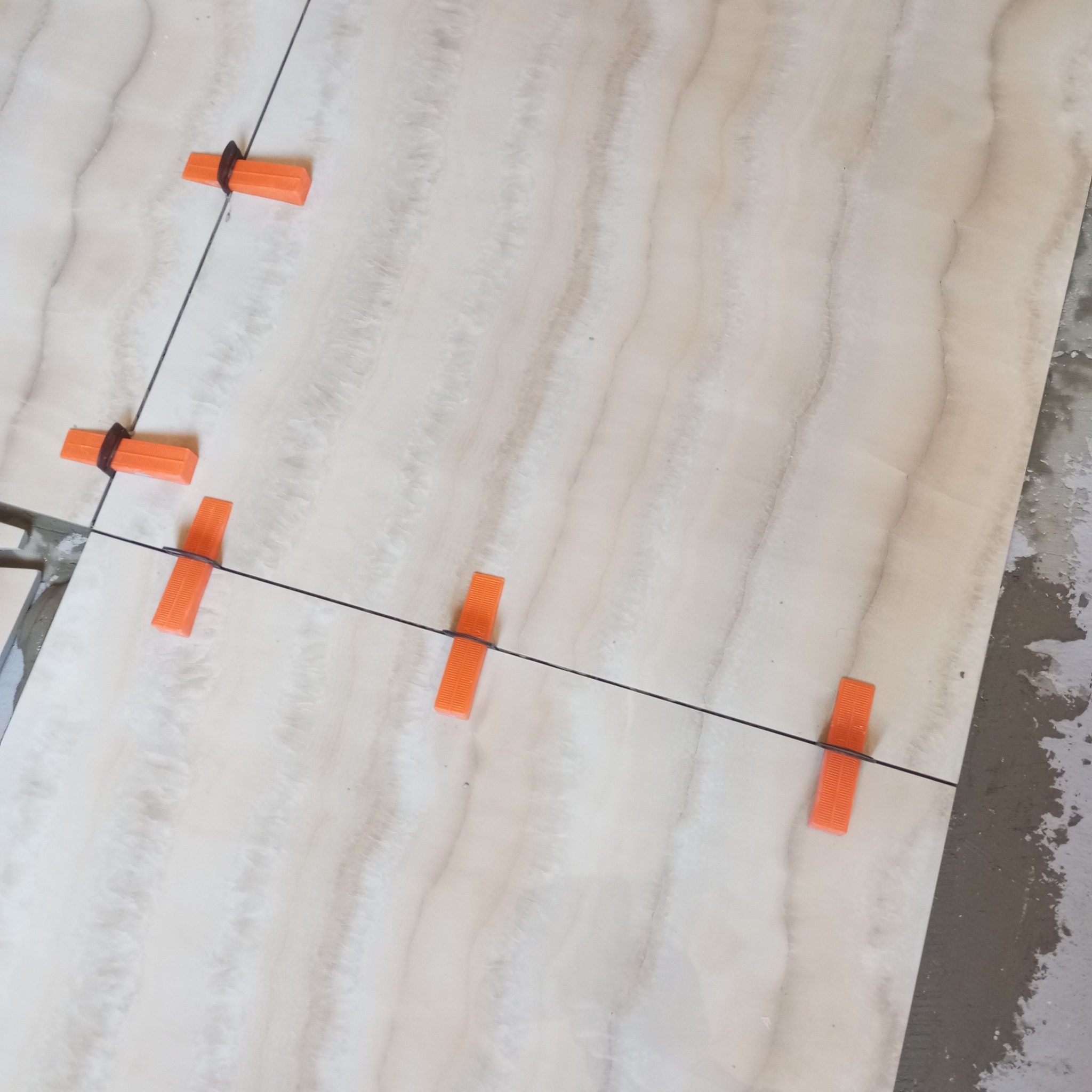 Układanie płytek imitujących marmur, z użyciem pomarańczowych klipsów poziomujących i czarnej żyłki, widok z góry, niedokończona podłoga.