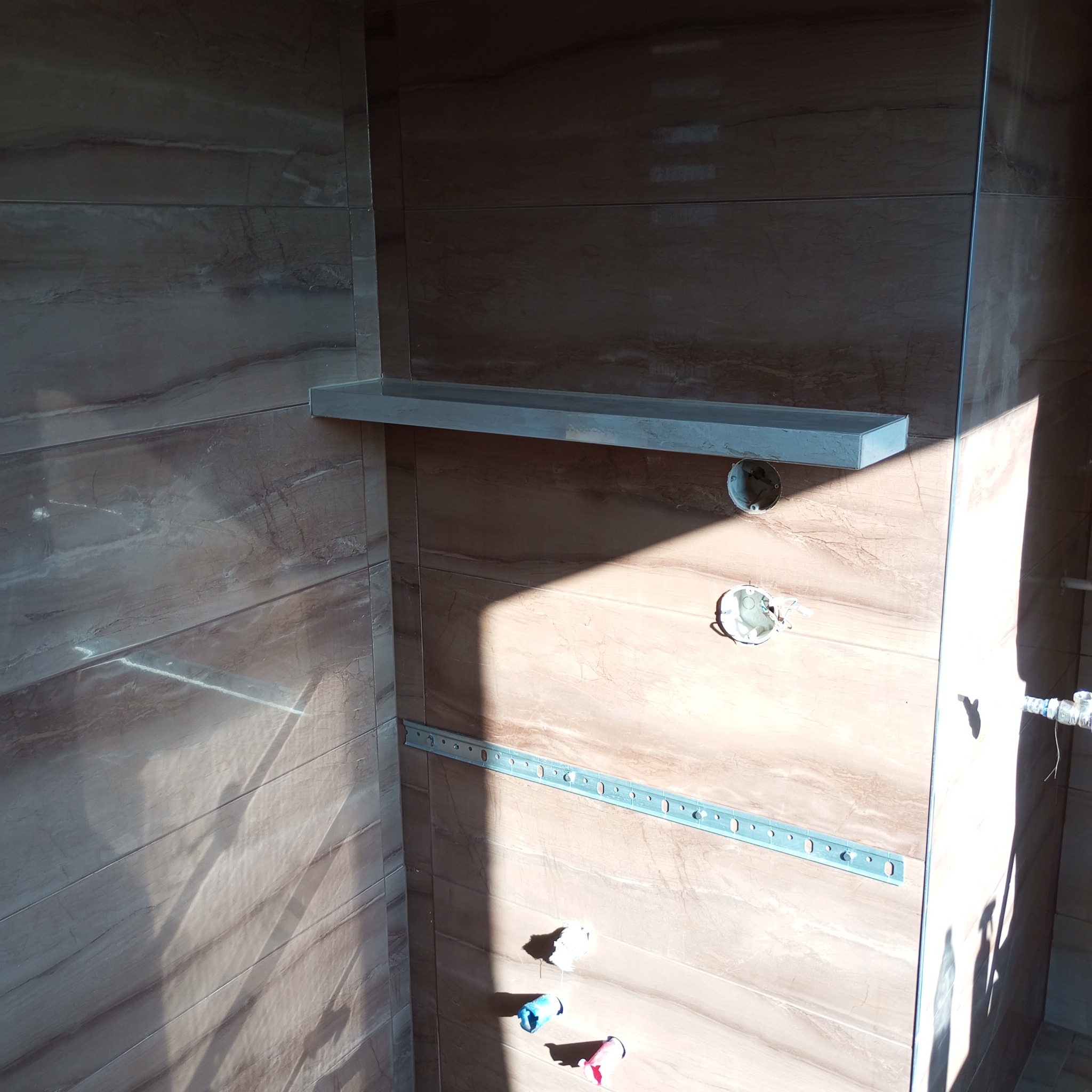 Ściana w łazience wyłożona brązowymi płytkami imitującymi marmur, widoczne puszki elektryczne, metalowy profil mocujący i półka w trakcie montażu, oświetlona silnym światłem słonecznym.