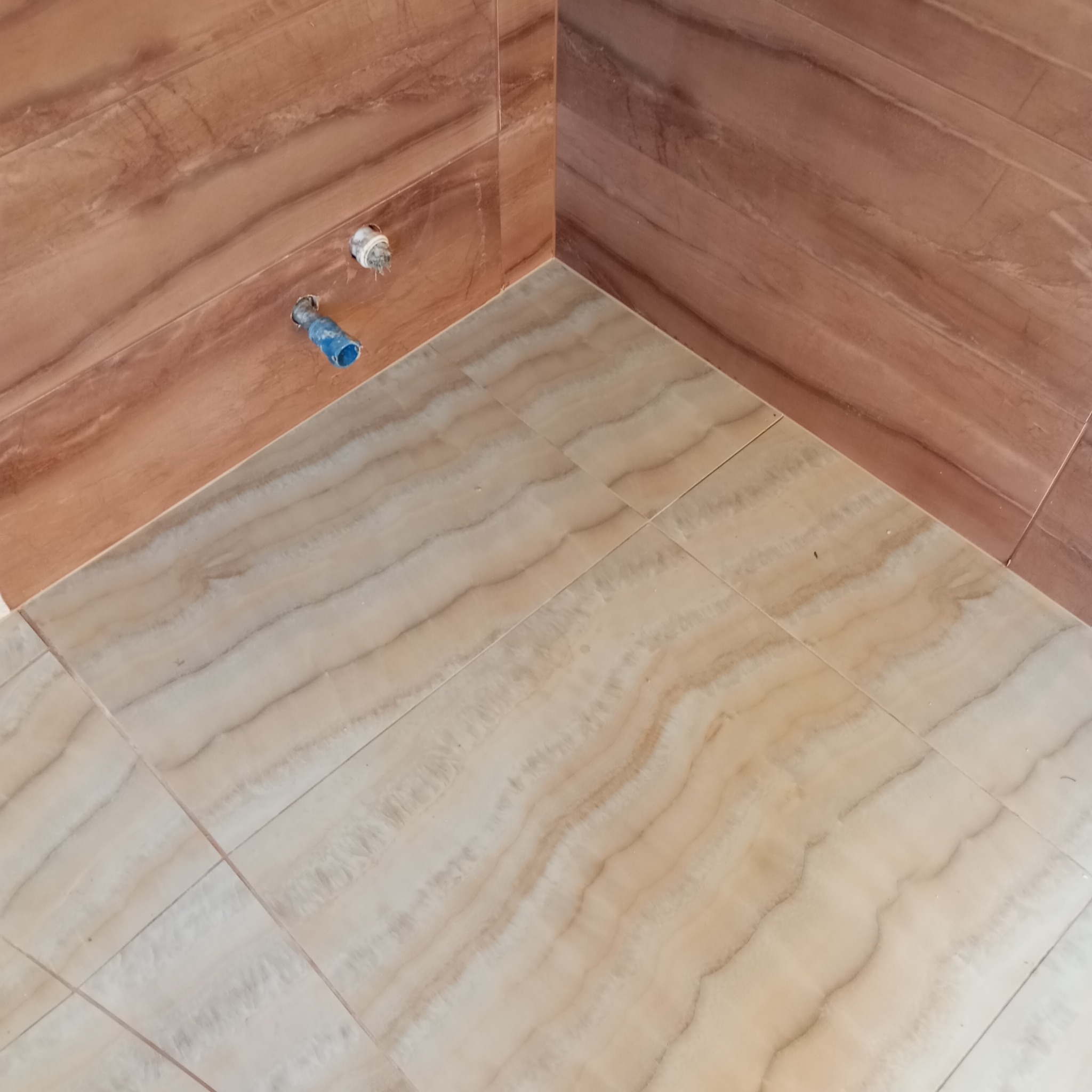 Wykończony narożnik łazienki z brązowymi płytkami ściennymi imitującymi drewno i beżowymi płytkami podłogowymi o wzorze marmuru, widoczne przyłącza hydrauliczne.