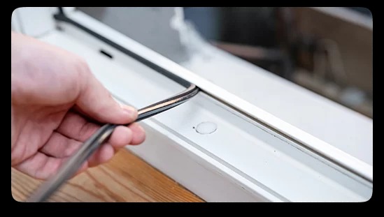 Ręka montuje uszczelkę okienną w białej ramie okna, widoczne drewniane podłoże.