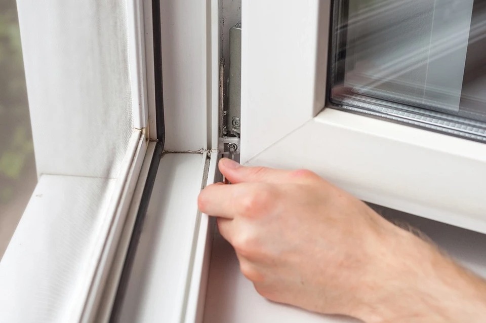 Regulacja okna PCV: dłoń dokręca śrubę w mechanizmie okiennym przy użyciu klucza imbusowego, widoczne uszczelki i zabrudzenia w narożnikach ramy.