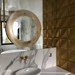 Nowoczesna łazienka z okrągłym lustrem w złotej ramie, białą umywalką nablatową, złotą armaturą i ścianą wykończoną złotymi panelami 3D.