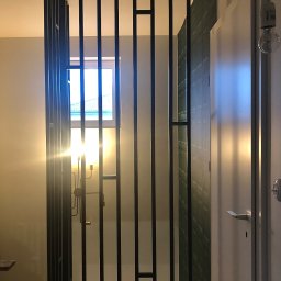 MASTER HOUSE - Nowoczesna, pionowa konstrukcja metalowa z czarnych profili, oddzielająca przestrzeń, z widokiem na okno i fragment ściany z wzorem oraz drzwi po prawej stronie.