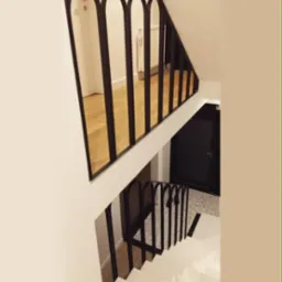 Schody z białymi stopniami i czarną, metalową balustradą z łukowatymi elementami, widok z góry, podłoga wyłożona parkietem i mozaiką.