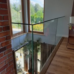 MASTER HOUSE - Wnętrze domu z widokiem na przeszkloną balustradę, przez którą widać roślinę doniczkową i rower, z ceglaną ścianą po lewej stronie i drewnianą podłogą po prawej.