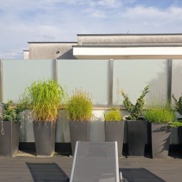 MASTER HOUSE - Nowoczesny taras z szarą podłogą, szklaną balustradą z metalowymi słupkami i rzędem wysokich, czarnych donic z różnorodną zielenią, częściowo zasłonięty leżakiem.