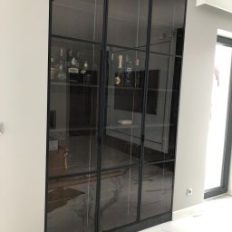 MASTER HOUSE - Nowoczesna, przeszklona witryna z czarnymi metalowymi ramami, prezentująca kolekcję alkoholi na marmurowej podłodze.