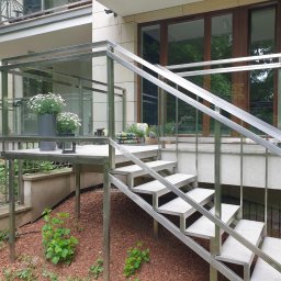 MASTER HOUSE - Zewnętrzne schody metalowe z balustradą nierdzewną prowadzące na podest z kwiatami w donicach, widok z dołu na tle budynku.