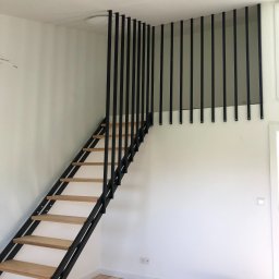 MASTER HOUSE - Metalowe schody z drewnianymi stopniami prowadzące na antresolę, zabezpieczone czarnymi, pionowymi prętami, widok z dołu, w tle białe ściany i podłoga w trakcie wykańczania.