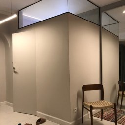 MASTER HOUSE - Nowoczesne wnętrze z zabudową antresoli nad ścianą, szklana konstrukcja z czarną ramą, drewniane krzesło z plecionym siedziskiem i porozrzucane kapcie na podłodze.