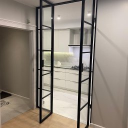 MASTER HOUSE - Dwuskrzydłowe, przeszklone drzwi z czarnymi, metalowymi szprosami oddzielające przedpokój z drewnianą podłogą od jasnej kuchni z marmurową podłogą i białymi meblami.