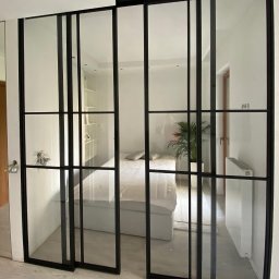 MASTER HOUSE - Przesuwne drzwi szklane z czarnymi szprosami, oddzielające sypialnię, zamontowane w domu w Nowej Wsi. Widoczne detale wykończenia podłogi i minimalistyczny wystrój wnętrza.