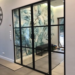 MASTER HOUSE - Nowoczesne, przeszklone drzwi wewnętrzne z czarnymi szprosami dzielącymi szkło na prostokątne pola, widok na korytarz z tapetą w kwiaty i ławką.