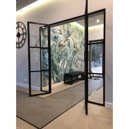 MASTER HOUSE - Nowoczesne, przeszklone drzwi wewnętrzne z czarnymi szprosami, otwarte na oścież, z widokiem na fototapetę z motywem kwiatowym i ławkę w tle, a także na zegar ścienny po lewej stronie.