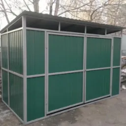 Zadaszona konstrukcja z zielonych paneli i szarej metalowej ramy, prawdopodobnie osłona na śmietniki, na tle drzew bez liści i fragmentu ogrodzenia.