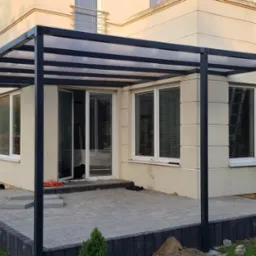 Nowoczesna, grafitowa pergola z przezroczystym zadaszeniem przylegająca do jasnego domu z oknami, widoczna drabina oparta o ścianę, świeżo ułożona kostka brukowa na tarasie.