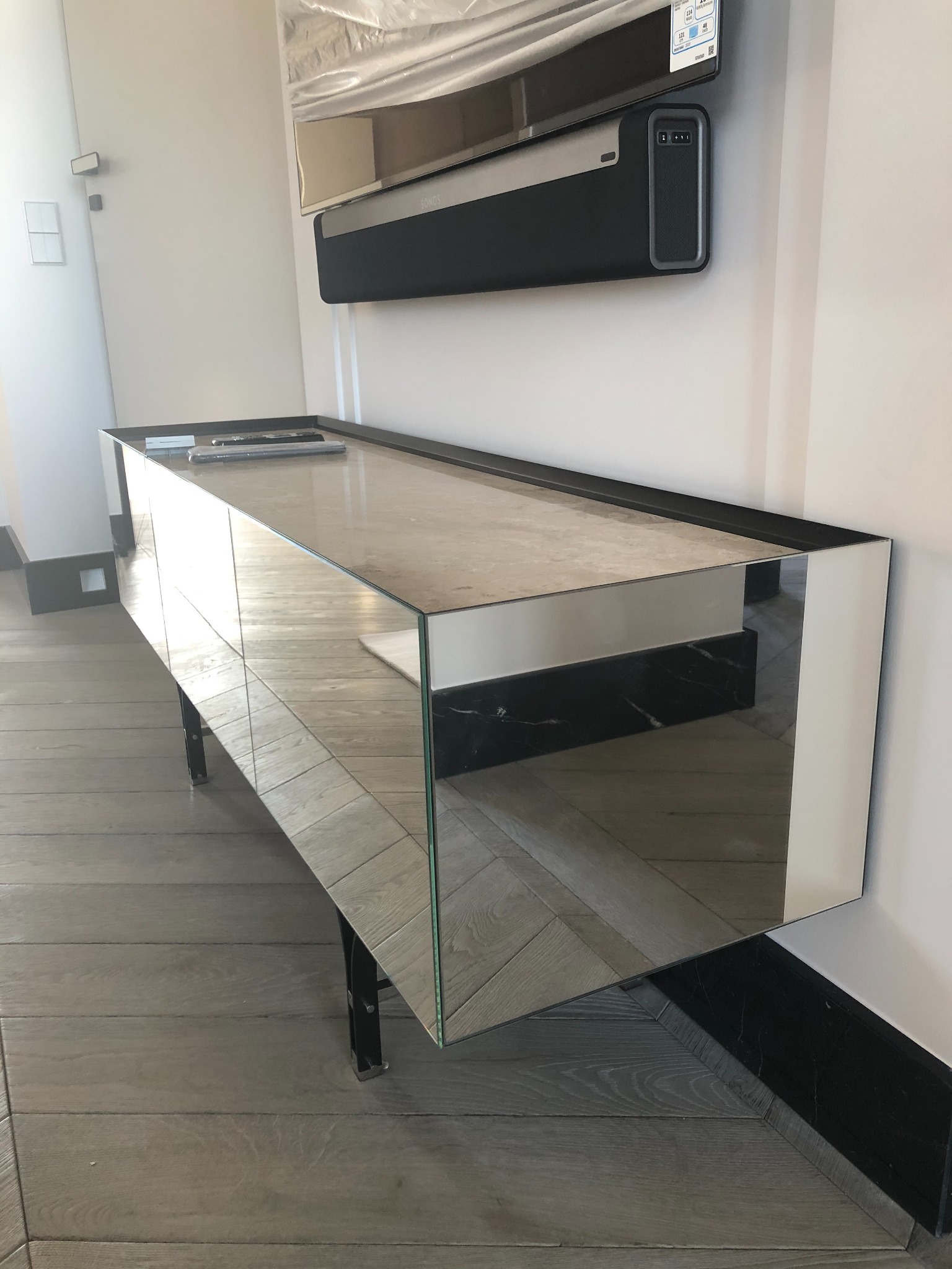 Nowoczesna szafka RTV z lustrzanymi frontami i marmurowym blatem, ustawiona na parkiecie w jodełkę, z widocznym telewizorem i soundbarem na ścianie.